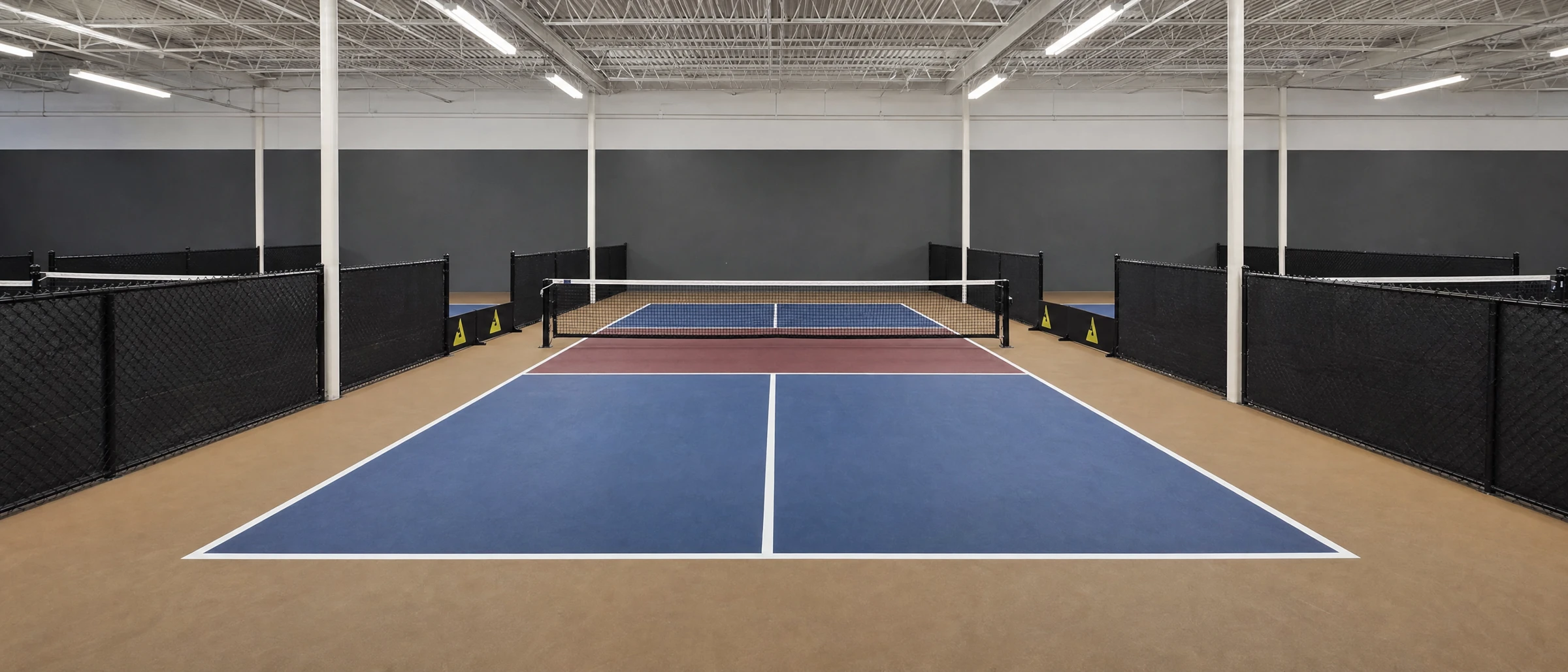Club 65 indoor pickleball court with tan-brown surrounds, blue service boxes, and burgundy kitchens